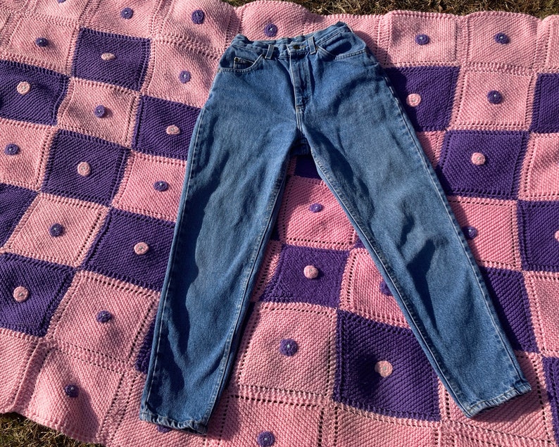 Puede incluir: Un par de pantalones vaqueros azules de mezclilla con cintura alta y pierna recta. Los pantalones vaqueros est&aacute;n colocados sobre una manta de crochet rosa y morada.