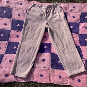 May include: A pair of light pink corduroy pants with a straight leg fit. The pants are laid flat on a pink and purple crocheted blanket.