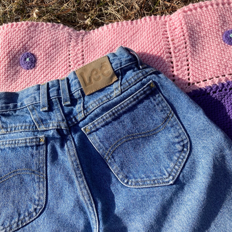 Puede incluir: Un par de pantalones vaqueros azules con un parche de cuero marr&oacute;n que dice "Lee" en la parte posterior. Los vaqueros tienen un bolsillo trasero con un dise&ntilde;o cosido.