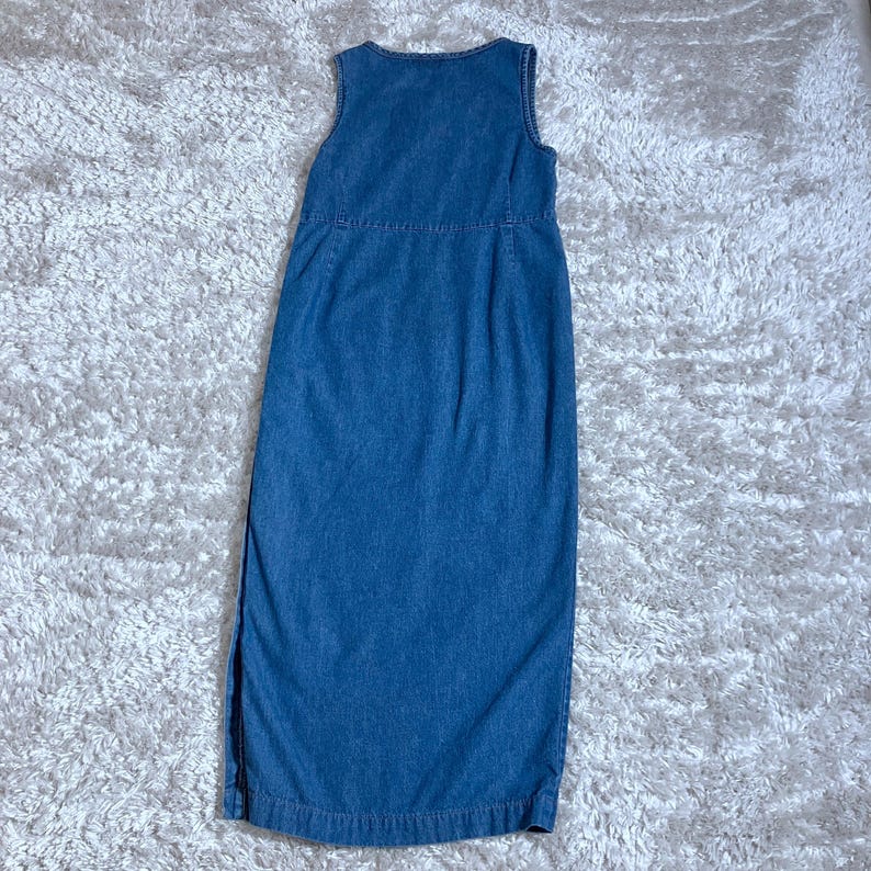 May include: A sleeveless, denim blue dress. The dress has a round neckline and a straight, floor-length silhouette. It features a side slit and a fitted bodice with a seam at the waist. The dress is made of a medium-weight denim fabric.