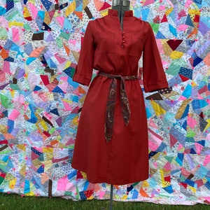 May include: A rust-colored vintage dress with a paisley patterned belt. The dress has a collared neckline and three-quarter sleeves. The dress is displayed on a mannequin against a colorful patchwork quilt background.