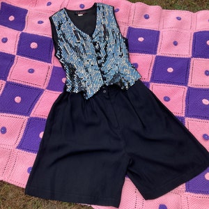 May include: A black sequined vest with a button closure and a pair of navy blue high-waisted shorts. The clothing is laid out on a pink and purple crocheted blanket.