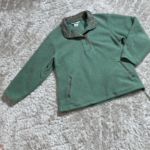 May include: A green fleece pullover with a button-up collar and a patterned collar lining. The pullover has a quarter-zip closure and two side pockets with zippers. The fleece is a solid green color, and the collar lining features a mix of brown, green, and tan.