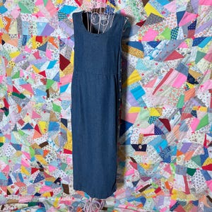 May include: A dark blue denim dress, sleeveless with a scoop neckline and fitted waist. The dress has a button closure down the side and a straight, ankle-length hem. It's displayed on a wire dress form against a colourful patchwork quilt background.