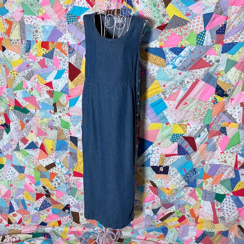 May include: A dark blue denim dress, sleeveless with a scoop neckline and fitted waist. The dress has a button closure down the side and a straight, ankle-length hem. It's displayed on a wire dress form against a colourful patchwork quilt background.