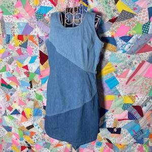 May include: A sleeveless denim dress in shades of light and medium blue. The dress features a unique diagonal panel design and a scoop neckline. The dress is displayed on a vintage metal dress form against a colorful patchwork quilt background.