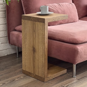 Pode incluir: Uma mesa lateral de madeira com acabamento natural está ao lado de um sofá rosa empoeirado. Uma xícara e um pires brancos estão sobre a mesa. Uma planta em vaso com um tronco torcido está à esquerda da mesa, contra uma parede de tijolos brancos.