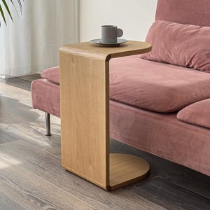 May include: A light-colored wooden side table with a rounded top and base, positioned next to a pink sofa. A gray mug and saucer sit on the table. The table is approximately 20 inches tall.