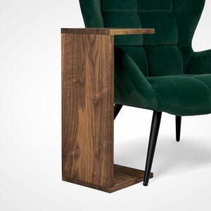 Peut inclure: Une table d'appoint en bois avec une finition brun foncé. La table a un plateau rectangulaire et une base carrée. La table est à côté d'un fauteuil en velours vert avec des pieds noirs.