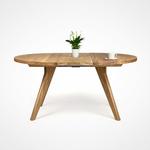 table with a massiv round top and four slanted legs, which taper downwards. On the top there is a flower in a white pot and a cup. the table has a unique grain pattern, typical of natural wood. the table is shown with one extension.