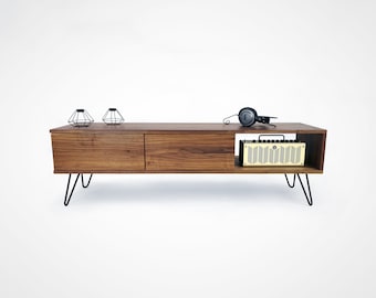 May include: A mid-century modern style walnut wood media console with two drawers and a shelf. The console has black hairpin legs and is decorated with two black wire geometric candle holders and a pair of black headphones.