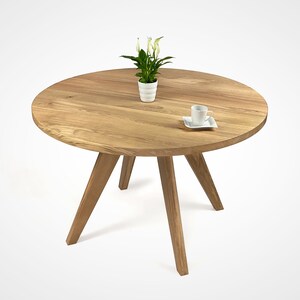 table with a massiv round top and four slanted legs, which taper downwards. On the top there is a flower in a white pot and a cup. the table has a unique grain pattern, typical of natural wood. the table is shown in a folded version.