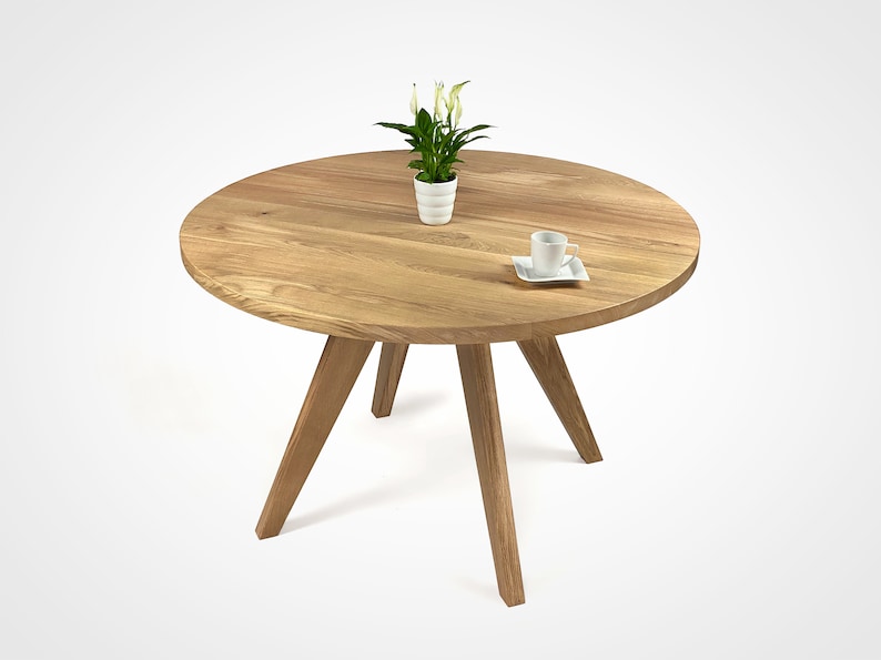 table with a massiv round top and four slanted legs, which taper downwards. On the top there is a flower in a white pot and a cup. the table has a unique grain pattern, typical of natural wood. the table is shown in a folded version.