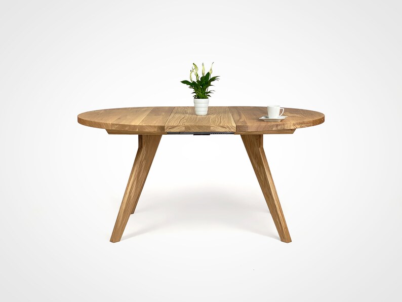 table with a massiv round top and four slanted legs, which taper downwards. On the top there is a flower in a white pot and a cup. the table has a unique grain pattern, typical of natural wood. the table is shown with one extension.