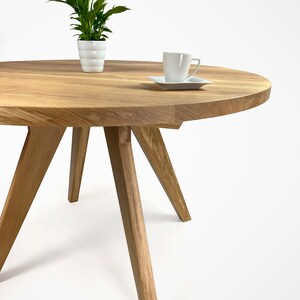 table with a massiv round top and four slanted legs, which taper downwards. On the top there is a flower in a white pot and a cup. the table has a unique grain pattern, typical of natural wood. the table is shown in a folded version.