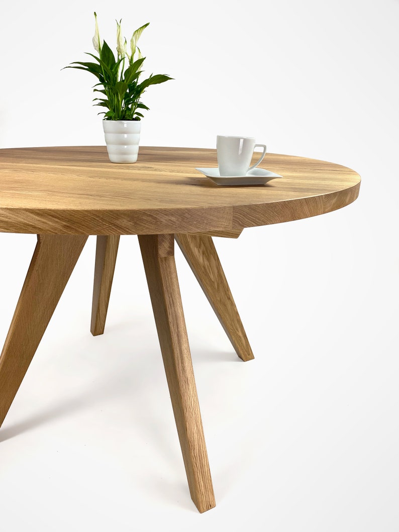 table with a massiv round top and four slanted legs, which taper downwards. On the top there is a flower in a white pot and a cup. the table has a unique grain pattern, typical of natural wood. the table is shown in a folded version.