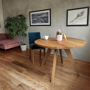 May include: A round wooden table with a natural finish and three legs. The table is set with a white cup and saucer and a small potted plant with pink flowers. A small wooden bird figurine is on the table.