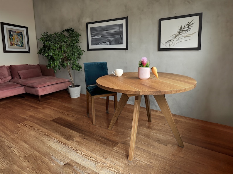 May include: A round wooden table with a natural finish and three legs. The table is set with a white cup and saucer and a small potted plant with pink flowers. A small wooden bird figurine is on the table.