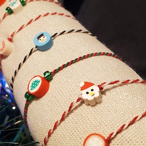 Peut inclure: Gros plan sur une décoration de Noël festive. Un tissu de couleur crème est enroulé de ficelle rouge, blanche et verte, orné de petites perles colorées sur le thème des fêtes. Les perles présentent des images de bonhommes de neige, de Père Noël et de sapins.