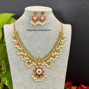 Op de afbeelding: Een goudkleurige ketting en oorbellen set. De ketting heeft een centrale hanger met een bordeauxrood en groen bloemmotief, omgeven door kleine parels en groene kralen. De oorbellen spiegelen het ontwerp. De tekst "Nsquare Collections" is zichtbaar.