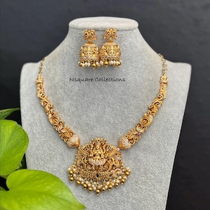 May include: A gold-toned necklace and earring set displayed on a mannequin. The necklace features an ornate pendant with a central figure and is adorned with small white beads. The matching earrings are bell-shaped with similar detailing. The text "Nsquare Collections" is visible.