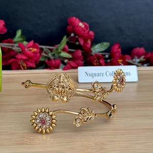 May include: Two gold-colored bangles with intricate floral and deity designs. One bangle features a central deity figure, while the other has a floral design with a central red stone. The background includes red flowers and the text "Nsquare Collins".