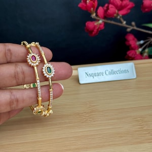 Puede incluir: Dos brazaletes dorados con una serie de pequeñas piedras claras y brillantes a lo largo de la banda. Cada brazalete está acentuado con piedras ovaladas rojas y verdes rodeadas por un halo de diminutas piedras claras. Un letrero blanco dice Nsquare Collections.