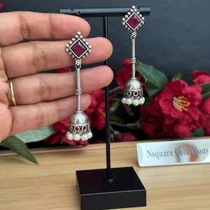 May include: Silver-coloured dangle earrings with a red square gemstone at the top and a bell-shaped bottom. The earrings feature a long, textured chain and small white pearls. The earrings are displayed on a black stand.