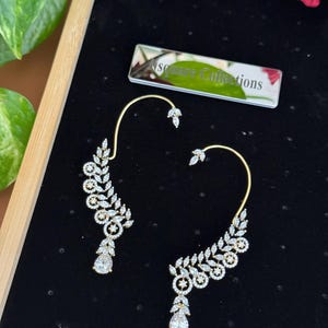 May include: A pair of gold-tone ear cuffs featuring a curved wire design adorned with clear marquise and round-cut stones in a leaf-like pattern, ending in a teardrop-shaped stone. A rectangular tag labeled Square Collections is included.