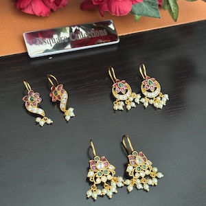 Puede incluir: Pendientes dorados con diseños florales, con piedras preciosas rojas, verdes y transparentes, y perlas blancas colgantes. Los pendientes se exhiben sobre una superficie oscura con un arreglo floral y un letrero metálico que dice "Nsquare Collections."