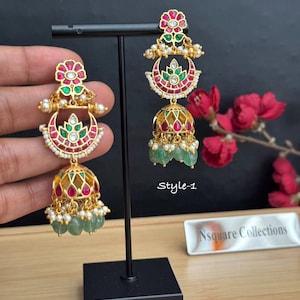 May include: A pair of gold-tone jhumka earrings featuring a floral top, a crescent moon center, and a bell-shaped base. The earrings are adorned with red and green stones, white pearls, and dangling green beads. The text Style-1 is visible.