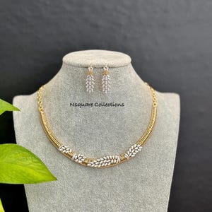Puede incluir: Un conjunto de collar y pendientes dorados exhibido en un maniquí. El collar presenta un diseño curvo con una sección central de piedras preciosas blancas. Los pendientes tienen forma de hoja colgante con piedras preciosas blancas. El texto "Nsquare Collections" es visible.