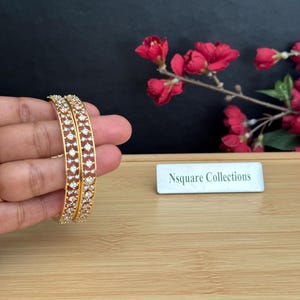 May include: Two gold-toned bangles featuring a lattice design encrusted with small, clear, round-cut stones. A rectangular white sign with the black text Nsquare Collections is positioned nearby on a light-coloured wooden surface.