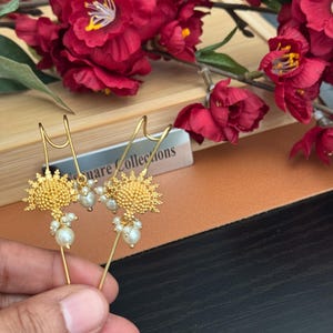 May include: A pair of gold-colored earrings with a sunburst design, adorned with small, white pearls. The earrings are held against a backdrop of red flowers and a wooden surface. The earrings are a unique jewelry item.