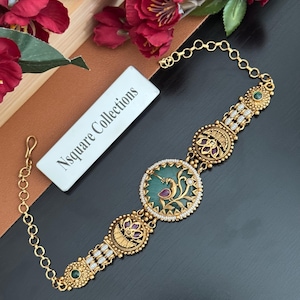 May include: A gold-toned bracelet featuring a central circular medallion with a green inlay and a peacock motif, flanked by smaller circular floral-patterned links with pink and green accents, finished with a chain and a white Nsquare Collections tag.