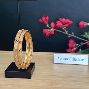 May include: Two gold-toned bangles featuring intricate textured patterns, small embedded red stones, and clear accents. The bangles are displayed on a black stand next to a white rectangular sign with the text Nsquare Collections printed in black.
