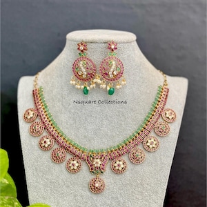 May include: A gold-toned necklace and earring set. The necklace features a series of circular pendants with red and white accents, connected by a green beaded strand. The earrings mirror the design, with dangling green beads and small pearls. The text "Nsquare Collections" is visible.