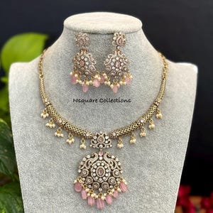 May include: A gold-toned necklace and earring set displayed on a mannequin. The jewelry features intricate detailing with clear and pink gemstones, and small pearl accents. The necklace has a large pendant with a floral design, and the earrings are matching. The text "Nsquare Collections" is visible.