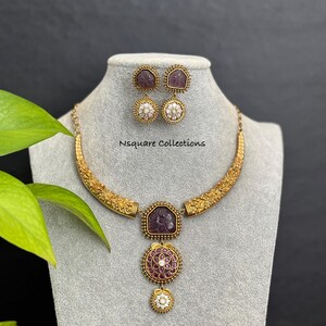 May include: A gold necklace and earring set on a grey mannequin. The necklace has a curved design with floral detailing and a purple gemstone pendant. The earrings have matching purple gemstones and white pearl accents. The text "Nsquare Collections" is visible.