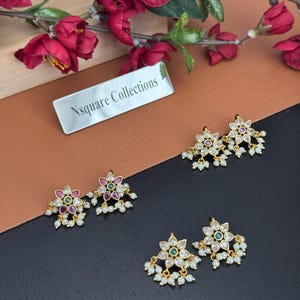 Op de afbeelding: Goudkleurige bloemen oorknopjes met kleurrijke edelstenen en bungelende witte parels. De oorbellen zijn gerangschikt op een bruin en zwart oppervlak, met een bordje met de tekst "Nsquare Collections" op de achtergrond.