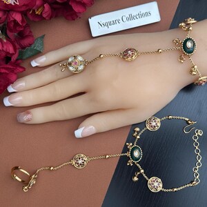 May include: Gold-coloured hand jewellery with intricate detailing. The set includes a ring connected to a bracelet by a delicate chain, adorned with red and green gemstones. The jewellery is displayed on a mannequin hand.