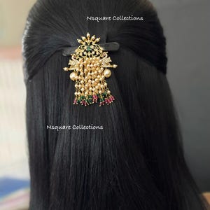 Peut inclure: Une barrette dorée avec un motif décoratif, des accents verts et rouges, des perles et des perles pendantes. La barrette est attachée à des cheveux noirs. Le texte "Nsquare Collections" est visible.