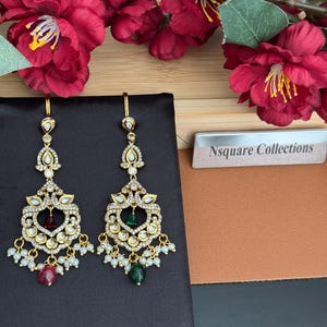 May include: Gold-coloured chandelier earrings with clear and red gemstones, and small white pearls. The earrings have a teardrop shape and are displayed on a black surface. The background includes red flowers and a sign that says "Nsquare Collections."