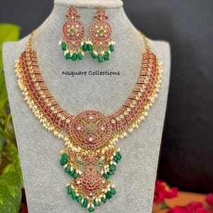 May include: A gold-toned necklace and earring set. The necklace features a central pendant with a circular design, surrounded by red and green gemstones, and dangling green beads. The earrings mirror the pendant design. The text "Nsquare Collections" is visible.