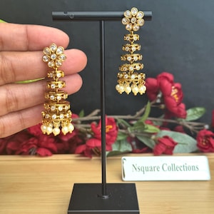 May include: Gold-toned chandelier earrings with a floral design at the top and multiple circular tiers. Each tier is adorned with clear gemstones and small, dangling pearls at the bottom. The earrings are displayed on a black stand against a dark background.