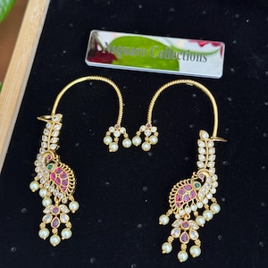 Pode incluir: Um par de brincos ear cuff dourados com um motivo de pavão adornado com cristais transparentes, pedras cor-de-rosa e pequenas pérolas brancas. O design inclui uma estrutura de arame curvo e elementos pendentes. Uma etiqueta com Nsquare Collections é visível.
