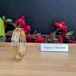 May include: Two gold-colored bangles with a geometric design and small, clear stones. The bangles are displayed on a wooden surface with red flowers and a sign that reads "Nsquare Collections."