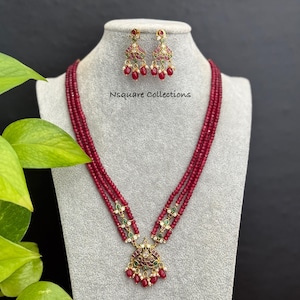 May include: A ruby red beaded necklace and matching earrings set. The necklace features multiple strands of small, round beads and a pendant with intricate gold detailing, red and green accents. The earrings mirror the pendant design. The text "Nsquare Collections" is visible.