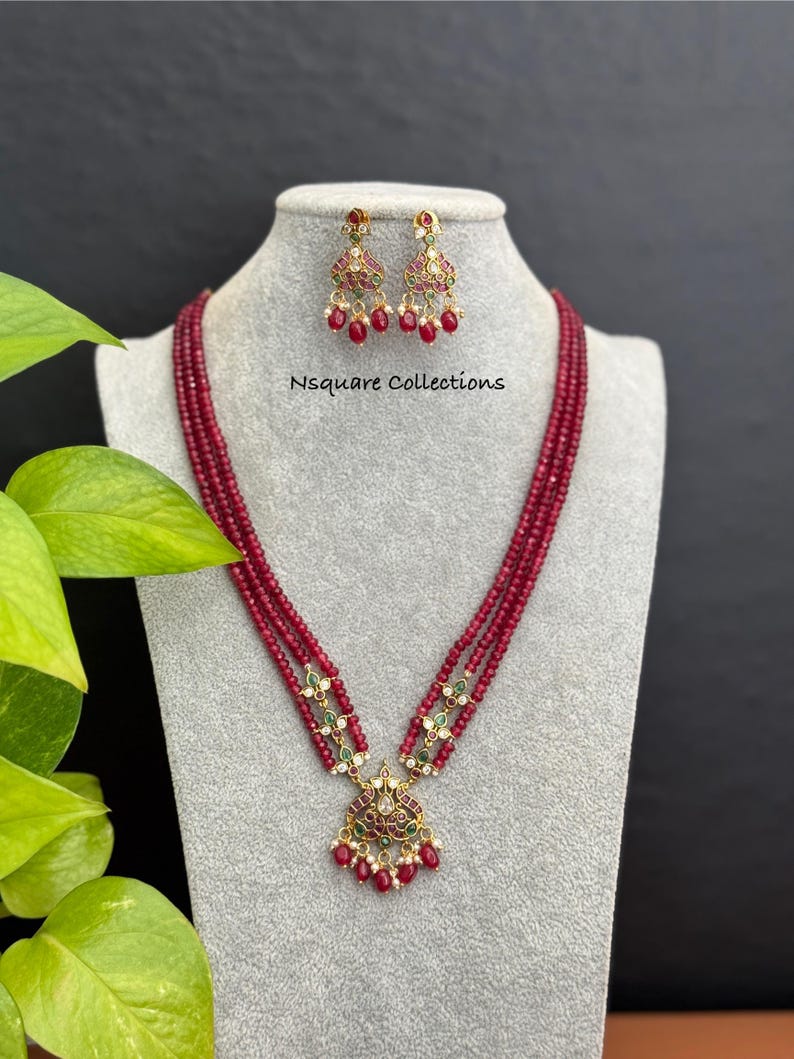 May include: A ruby red beaded necklace and matching earrings set. The necklace features multiple strands of small, round beads and a pendant with intricate gold detailing, red and green accents. The earrings mirror the pendant design. The text "Nsquare Collections" is visible.