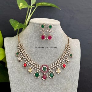 May include: A necklace and earring set featuring clear crystals, oval-shaped green, red, and yellow stones set in a metallic frame. The necklace has a central pendant with a green stone surrounded by red and clear crystals. Text reads Nsquare Collections.
